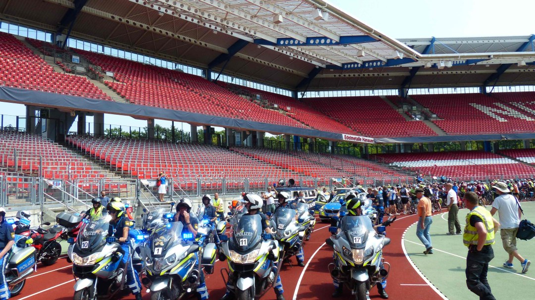 Im Max-Morlock-Stadion stehen auf der Tartanbahn vor der Sitztribüne  viele Polizisten auf Motorrädern. Dahinter stehen die Teilnehmenden der BR-Radltour mit ihren Fahrrädern.