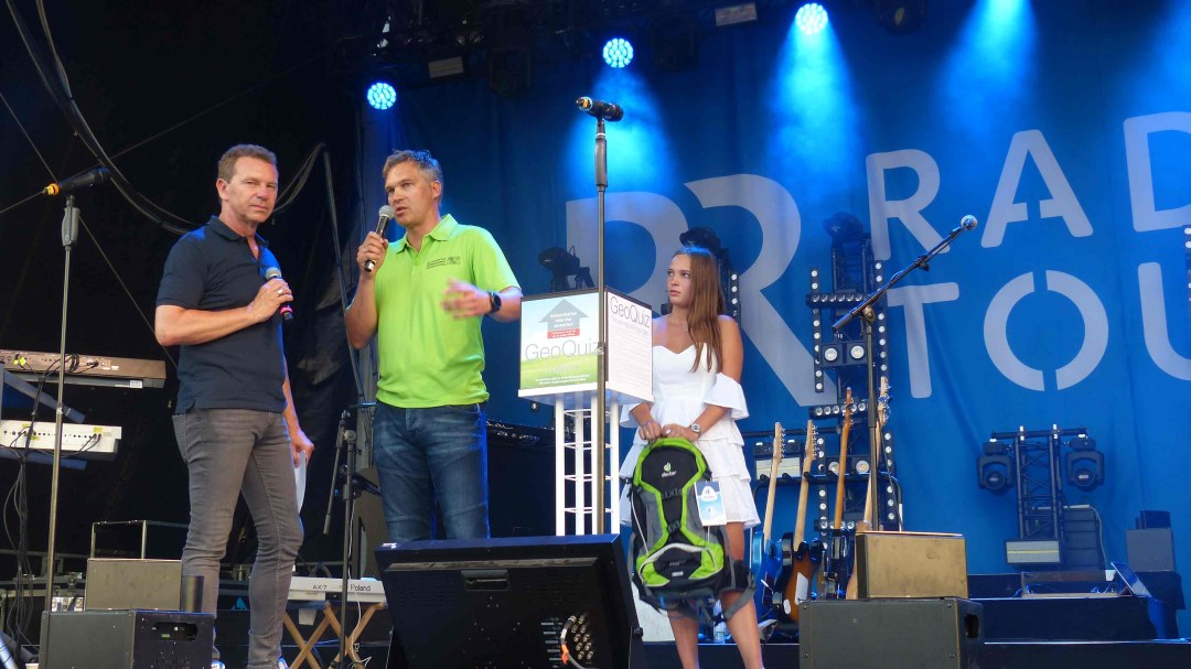 LDBV-Präsident Wolfgang Bauer und Moderator Roman Roell stehen auf der BR-Radltour-Bühne und reden in Mikrophone. Neben ihnen steht eine junge Frau, die einen Rucksack in der Hand hält.