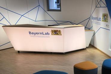 Im Eingangsbereich eines BayernLabs steht ein Counter aus mattweißem Plexiglas der von innen beleuchtet wird. Darauf steht 